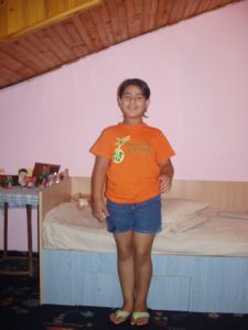 Girl in orange shirt standing by bed.